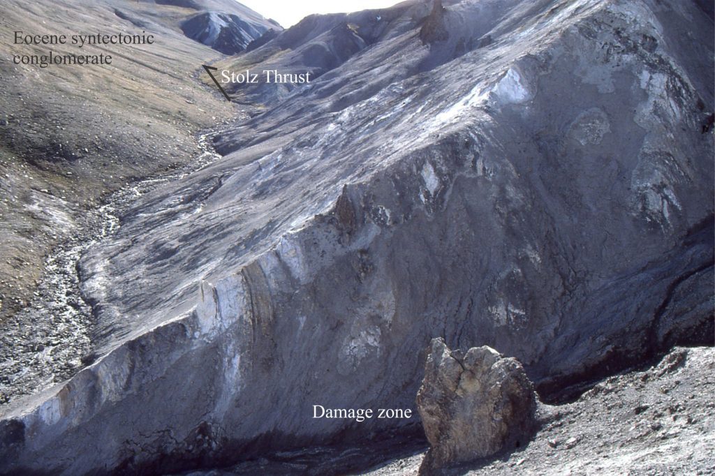 Damage zone in the hanging wall of Stolz Thrust (Eocene); Footwall contains Middle Eocene syntectonic conglomerate, Axel Heiberg Island, Canadian Arctic. The zone consists of open drag folds in interbedded sandstone (white) and shale, and significant shearing that has obliterated some of the original bedding. The bulk permeability in this zone is low. The zone in this view is about 20 m wide.