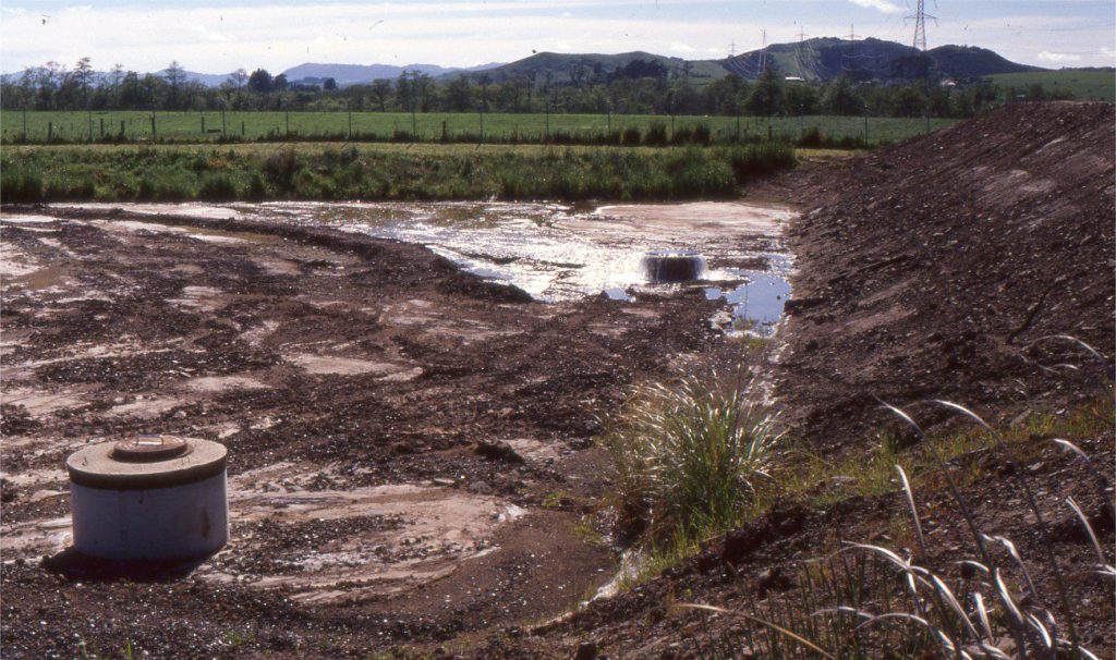 Anthropogenic colluvium (Category 5) created by excavation and redistribution of Late Pleistocene-Holocene flood-plain sand and gravel (flanking Waikato River). The original textural and stratigraphic characteristics of the flood plain deposits have been destroyed and replaced by a non-sorted, haphazardly stratified to non-stratified colluvium that is periodically redistributed by sheet flood runoff. The well-head right-centre is an artesian flowing outlet connected to the reservoir behind the embankment. Located near Huntly, New Zealand.