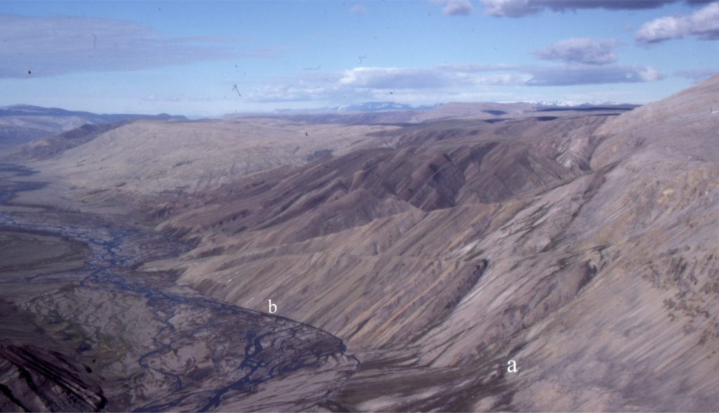 This view shows the transition from colluvium to alluvium via different landforms. The hill slopes flanking the braided river (centre-right) are underlain by steep dip-slopes of interbedded Paleocene-Eocene sandstone and shale. Small talus fans are accumulating at the base of these slopes. The colluvium is a mix of Category 1 and 2 types – mass wasting and slope wash (i.e., Type 3 – hybrid), the latter forming during the Arctic spring thaw. In the foreground (a) the talus fans merge at an abrupt break in slope into small alluvial fans. At (b) the talus fans merge abruptly with the braided river channels. Sediment distribution beyond the confines of the colluvium at (a) and (b) occurs primarily by channelized flow. Mt. Moore area, southern Ellesmere Island.